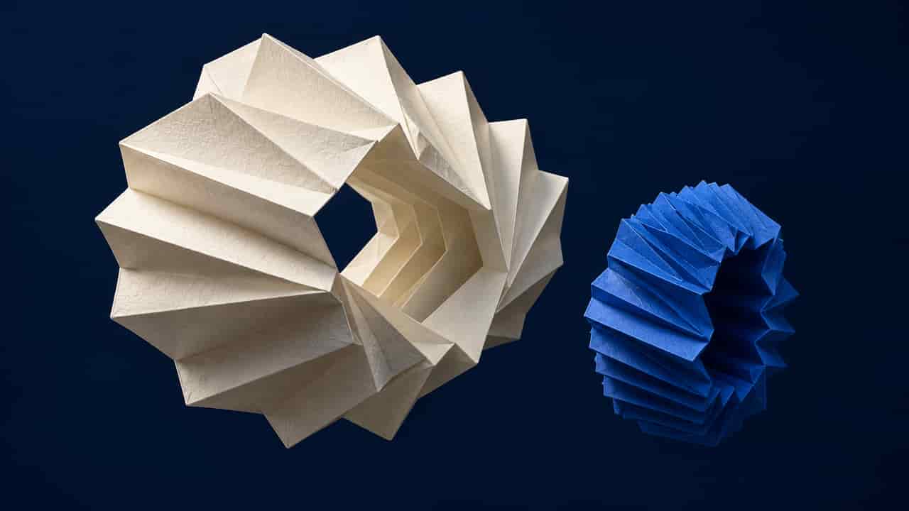 Two suspended folded-paper rings, one broad and ivory and one compact and cobalt blue, have differently shaped central openings against a dark navy background.