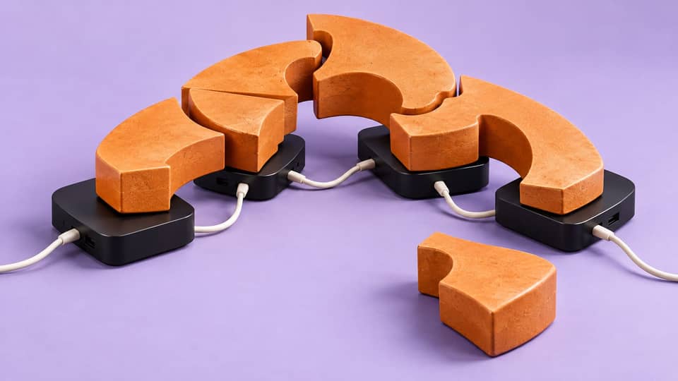 Orange interlocking sculpture segments sit on connected black TV boxes, with one loose segment illustrating that working parts do not establish a complete system.